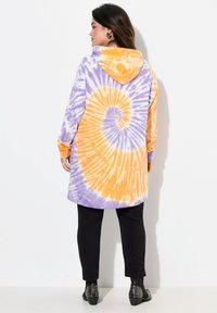 Abbigliamento a maniche lunghe con cappuccio, caratterizzato da un motivo a spirale in tie-dye viola e arancione, realizzato in tessuto leggero e con una vestibilità comoda. Abbinato a pantaloni neri a sigaretta.
