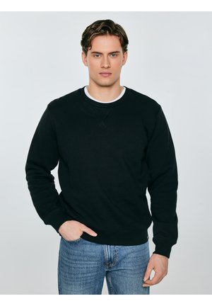 Młody mężczyzna o falowanych brązowych włosach, ubrany w czarną bluzę z okrągłym dekoltem i niebieskie dżinsy, stojący z jedną ręką w kieszeni na jednolitym tle.