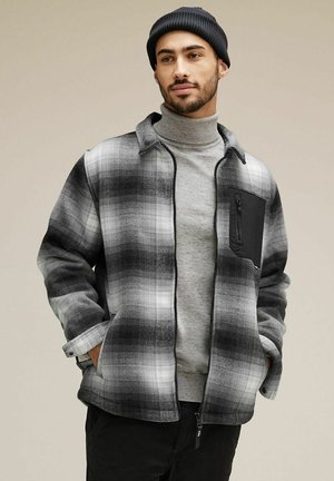 Sort og grå ternet jakke med lynlås, stående krave og en sort lynlåslomme på brystet. Stoffet ser blødt og struktureret ud.