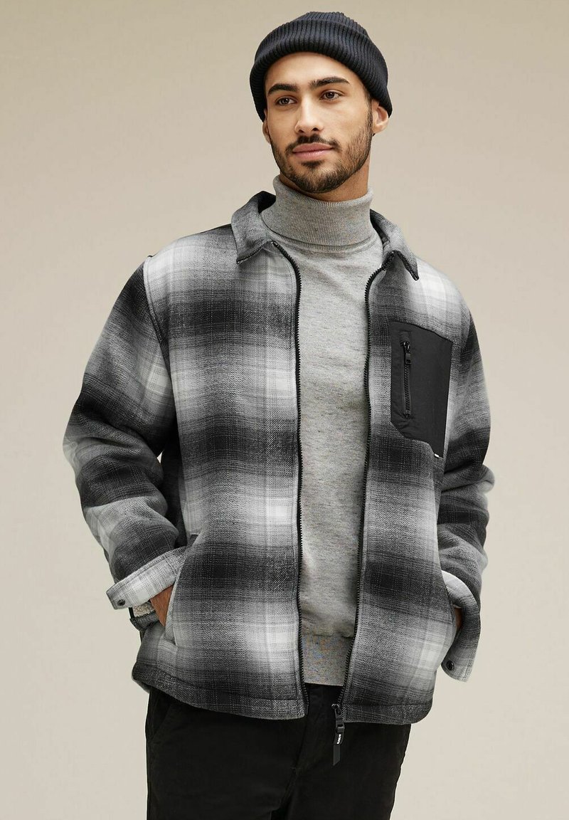 Svart och grå rutigt jacka med dragkedja, ståkrage och en svart dragkedjeficka på bröstet. Tyget verkar mjukt och texturerat.