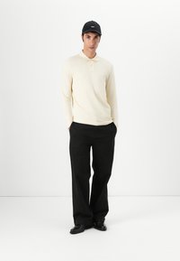 Polo à manches longues jaune pâle avec col et poche, associé à un pantalon noir ample et des chaussures noires, le tout complété par une casquette noire.