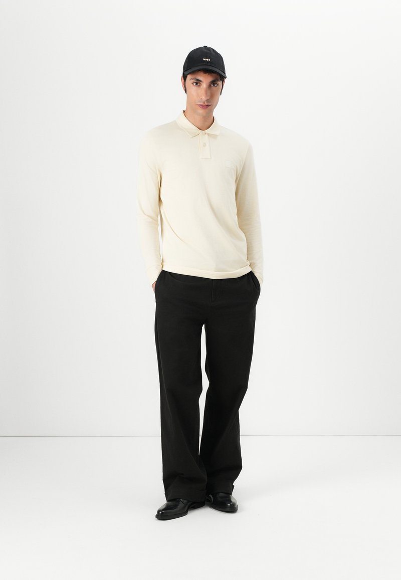 Langärmliges hellgelbes Poloshirt mit Kragen und Tasche, kombiniert mit weiten schwarzen Hosen und schwarzen Schuhen, gekrönt von einer schwarzen Kappe.
