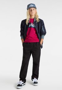 Laivasto sininen pommittaja takki, fuksia T-paita jossa on "VANS" graafinen painatus, mustat housut ja mustat/valkoiset lenkkarit. Harmaa lippis jossa on logoyksityiskohta. Pitkä vaalea tukka.
