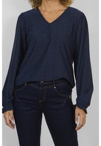 Blouse bleu marine à manches longues avec un col en V, présentant des motifs de broderie ajourée et des poignets élastiqués, associée à un jean en denim foncé.