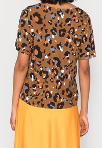 Chemise brune à manches courtes présentant un motif léopard en noir, orange et bleu. Le tissu semble lisse avec une coupe décontractée.