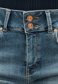 Κοντινό πλάνο της ζώνης παντελονιού από μπλε τζιν, που εμφανίζει διπλά χαλκινα κουμπιά, θηλιές για ζώνη και λεπτομερή πορτοκαλί ραφές.