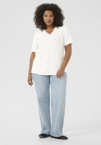 Haut blanc à manches courtes avec un détail en dentelle au niveau du col, associé à un jean droit bleu clair. Le mannequin se tient sur un fond uni.