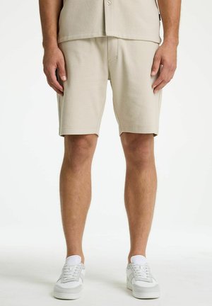 Mann trägt beige strukturierte Shorts, weiße Sneaker und ein dazu passendes beiges Hemd mit Knöpfen, steht vor weißem Hintergrund.