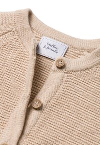 Beige Strickjacke mit Rundhalsausschnitt, Holzknebel und einem aufgenähten Etikett mit der Aufschrift "Stellou & friends." Sichtbares detailreiches Gewebe.