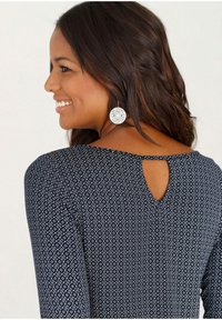 Marineblaues Kleid mit weißem Diamantmuster, ausgestattet mit einem Schlüsselrücken-Detail und langen Ärmeln. Das Modell trägt große runde Ohrringe.