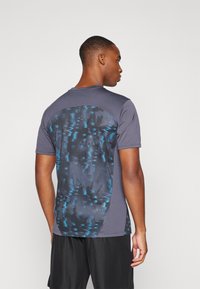 Camiseta deportiva gris con un panel trasero texturizado de patrón azul, mangas cortas y un escote redondo. También se ven pantalones cortos negros.