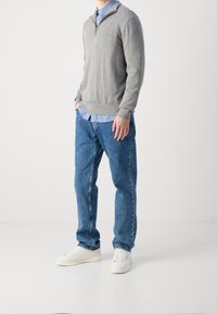 Maglione grigio con zip sopra una camicia azzurra a colletto, abbinato a jeans di denim blu e sneaker bianche. Look casual, texture morbida.