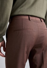 Braune maßgeschneiderte Hosen mit einer glatten Textur, ausgestattet mit zwei Gesäßtaschen und einem subtil gewebten Stoffdesign, kombiniert mit einem grünen Oberteil.