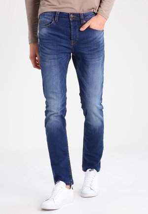 Men's blue denim jeans featuring a slim fit, light fading, front pockets, and a classic button fly, paired with white sneakers.
