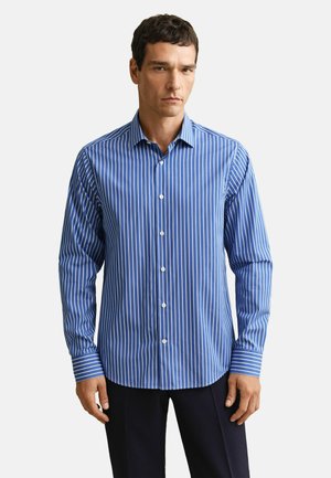 Man draagt een blauw-wit verticaal gestreept overhemd met lange mouwen en zwarte broek, staand tegen een effen achtergrond.