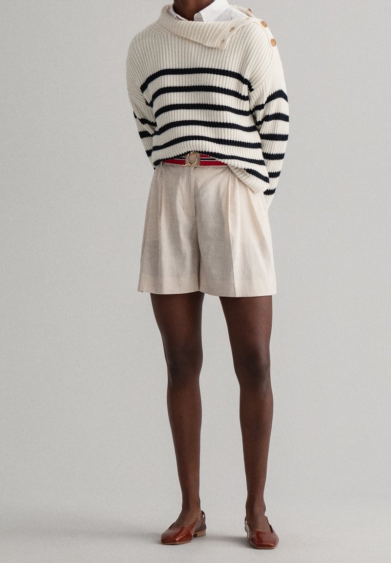 Pull en maille crème avec rayures bleu marine, coupe oversize, détail de boutons sur l'épaule. Shorts beige avec plis, chaussures plates marron.