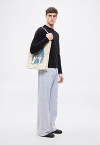 Τσάντα ώμου από καμβά με γραφικό σχέδιο σε κρεμ φόντο. Το μοντέλο φοράει μαύρο πουλόβερ και ανοιχτό μπλε τζιν με φαρδιά πόδια.