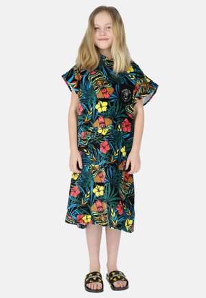 Młoda dziewczyna o blond włosach, ubrana w kolorową sukienkę z tropikalnym wzorem oraz czarne sandały typu slide na białym tle.