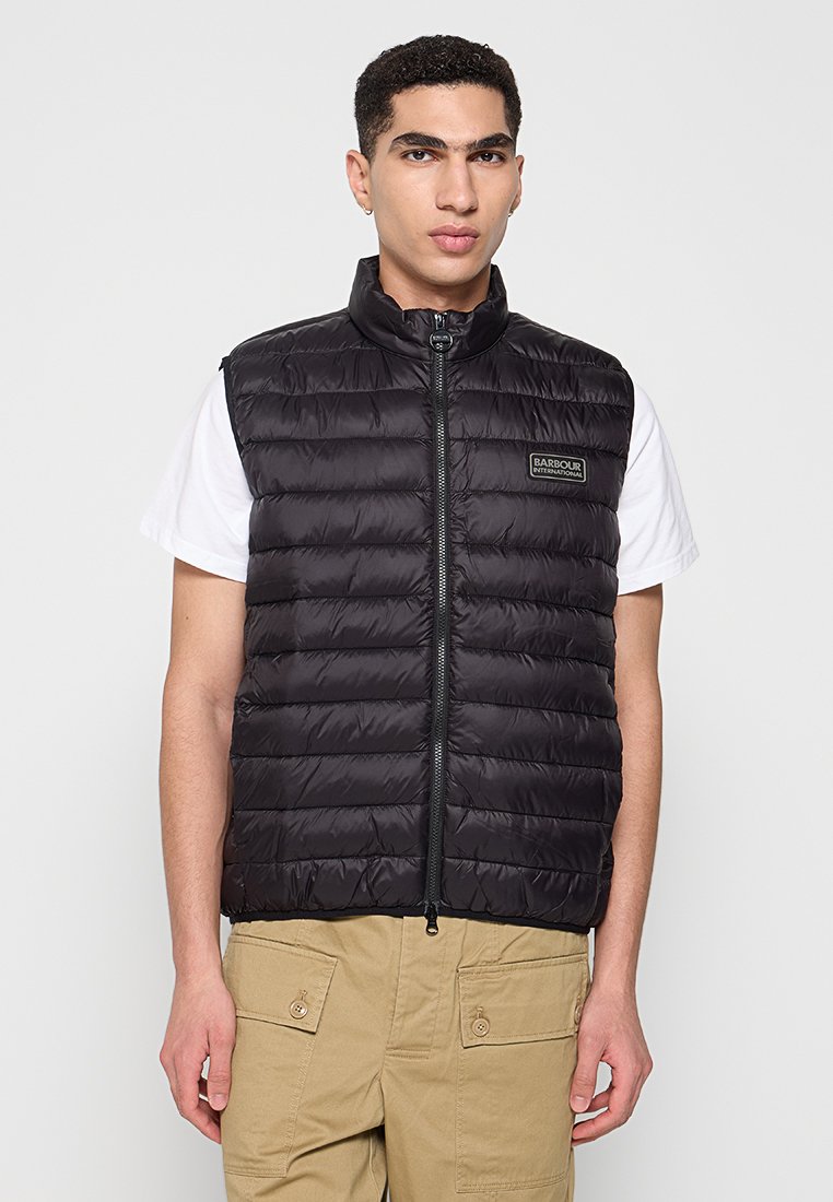 Schwarze Weste aus Daunen mit hohem Kragen, Reißverschlussfront und horizontalen Nähten. Verfügt über ein Logo-Patch auf der linken Brust. Tragen über einem weißen T-Shirt.