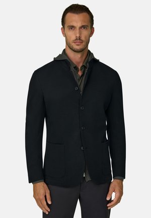 Homme portant un blazer noir boutonné sur une chemise col vert foncé, debout devant un fond gris clair uni.