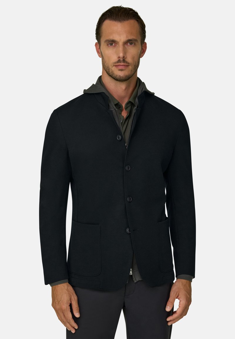 Homme portant un blazer noir boutonné sur une chemise col vert foncé, debout devant un fond gris clair uni.