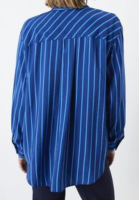 Persona che indossa una camicia blu di taglio ampio con strisce verticali e diagonali azzurre, vista da dietro su uno sfondo semplice.
