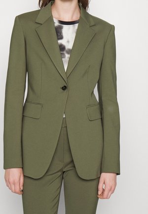 Donna che indossa una giacca da completo verde oliva su misura e pantaloni abbinati sopra una camicia fantasia bianca e nera.