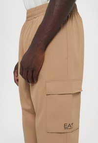 Pantalons cargo beiges avec une taille élastique, une poche latérale et un logo brodé sur la cuisse. Tissu lisse avec une coupe ample.