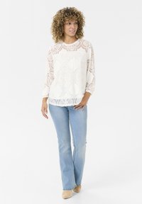 Top bianco in pizzo con motivi floreali, maniche a tre quarti e orlo scallop, abbinato a jeans azzurri a zampa d'elefante e scarpe beige.