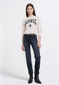 Béžová mikina s dlouhým rukávem s nápisem "ICONIC" tištěným černě, doplněná tmavými džínami a černými kotníkovými botami. Jednoduchý design, volný střih.