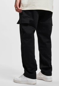Pantalons cargo noirs avec de multiples poches, présentant une coupe décontractée et une jambe droite. Fabriqués en tissu durable, associés à des baskets blanches.