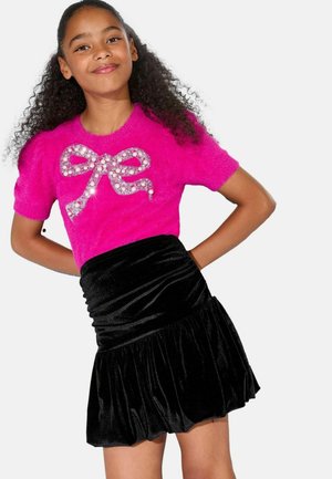 Chica con cabello rizado que lleva un suéter esponjoso de color rosa brillante con un lazo de perlas y lentejuelas, y una falda negra de terciopelo fruncida.
