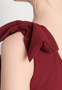Robe bordeaux en tissu doux et texturé, avec un grand nœud à l'épaule et une silhouette ajustée.