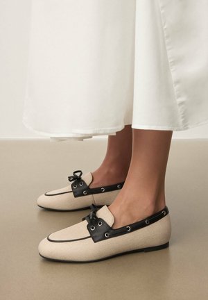 Beige woven loafers with a black leather accent and eyelets. Features a small bow on the front, flat sole, and rounded toe design.
