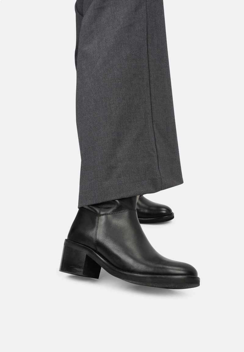 Bottines en cuir noir à talons carrés portées sous un pantalon large gris foncé sur fond blanc.