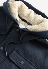 Navy blue jacket with a textured outer fabric, fluffy cream lining, a hood, a front pocket, and black snap buttons.