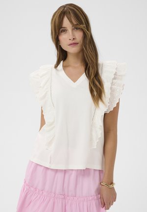 Femme aux longs cheveux bruns portant un chemisier blanc sans manches à volants et une jupe rose à étages, accessoirisée d'un bracelet en or.