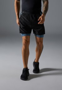 Svarte sportshorts med blå detaljer, med elastisk liv. Modellen kombinerer dem med svarte sportssko.