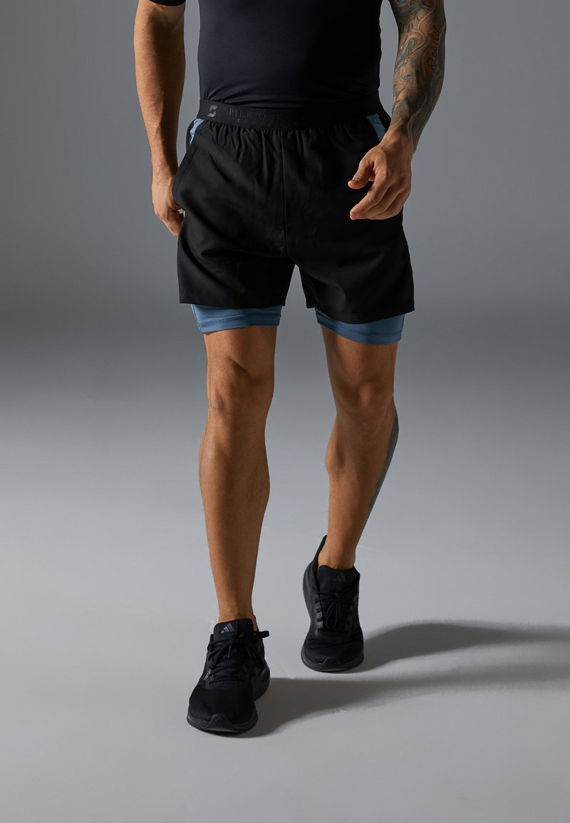 Svarte sportshorts med blå detaljer, med elastisk liv. Modellen kombinerer dem med svarte sportssko.
