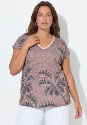 Vrouw met golvend bruin haar draagt een mauve V-hals top met abstracte zwarte en gouden patronen en witte broek, staand tegen een witte achtergrond.