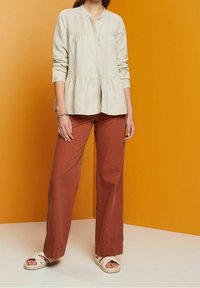Blusa crema de botones con dobladillo peplum, combinada con pantalones de pierna ancha color óxido y sandalias blancas con tiras, todo sobre un fondo naranja brillante.