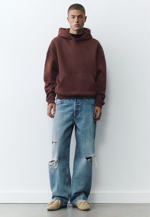 Giovane uomo che indossa una felpa marrone con cappuccio, jeans blu larghi e strappati e sneakers beige, in piedi contro un muro bianco semplice.