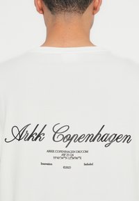 Bílá bavlněná tričko s černým kurzívním logem "Arkk Copenhagen" na zádech, pod logem se nachází další text a souřadnice.