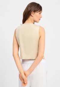 Beige, kurz geschnittenes Tanktop mit glatter Textur und Rundhalsausschnitt, ohne Ärmel und mit sauberem Saum, kombiniert mit hell gefärbten Hosen.