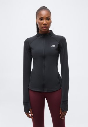 Vrouw met donker haar in een paardenstaart, die een zwart sportjack met rits en wijnrode legging draagt, staande tegen een effen lichte achtergrond.