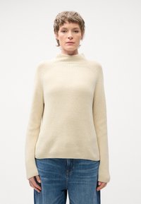 Beige, bordázott kötött pulóver magas nyakkal és hosszú ujjakkal, kék farmernadrággal párosítva, laza szabással és texturált felülettel.