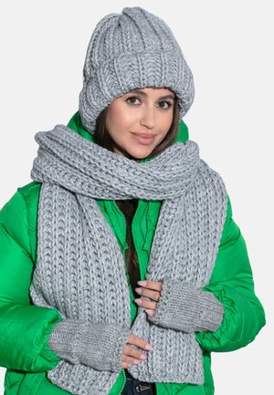 Szary zestaw czapki beanie i szalika z ribbowanym wzorem, w połączeniu z zieloną pikowaną kurtką. Rękawiczki bez palców wykonane z pasującego szarego materiału dzianego.
