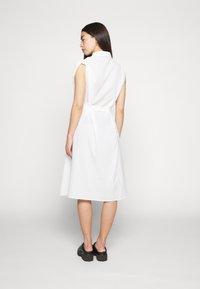 Vestido branco, sem mangas, com colarinho, cintura ajustada e saia até ao joelho. Tecido texturizado com um padrão listrado subtil. Calçado preto.