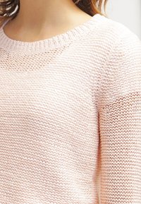 Pull en maille rose clair avec un col rond, un motif texturé et des finitions côtelées au niveau du col et des poignets.
