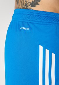 Blå atletiske shorts lavet af glat stof med elastisk talje, med "AEROREADY" logo og tre lodrette hvide striber.